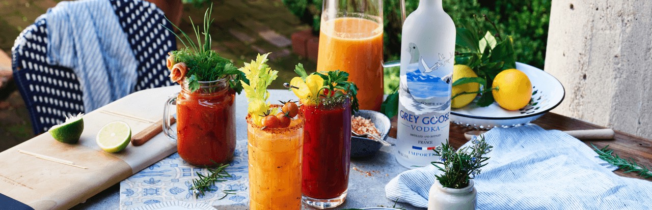Flasche Grey Goose Vodka mit frisch zubereiteten Bloody Mary Cocktails auf einem stilvoll gedeckten Gartentisch, umgeben von frischen Zutaten wie Zitrone, Kräutern und Saft. Ideal für sommerliche Cocktail-Momente.