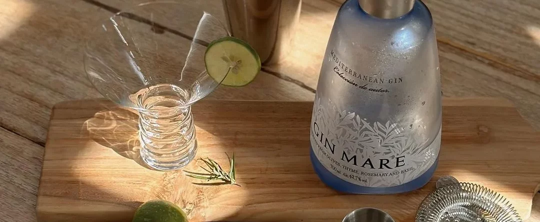 Gin Mare – mediterraner Premium-Gin mit Aromen von Oliven, Thymian, Rosmarin und Basilikum, servierbereit mit Shaker, Barzubehör, Limetten und Cocktailglas auf einem rustikalen Holztisch.